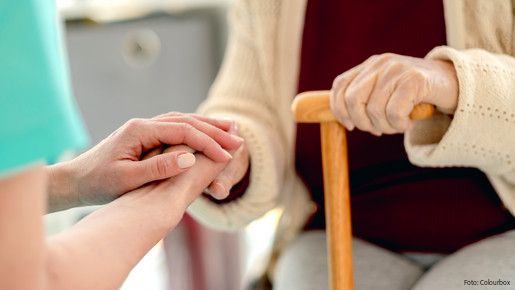 Das Foto zeigt die Hände einer Pflegerin, die die Hände einer Seniorin mit einem Stock halten. Es handelt sich um eine Nahaufnahme.