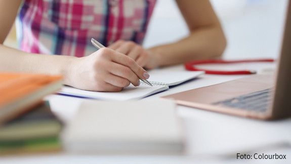 Initiative kulturelle Integration - Schreib für Hanau! Das Foto zeigt die Hand einer Person, die mit einem Stift auf einem Block schreibt. Rechts im Bild steht ein aufgeklapper Laptop.