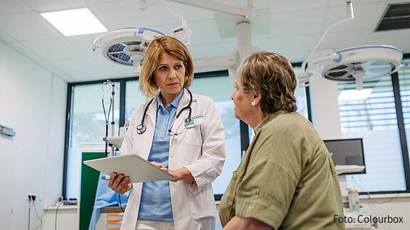 Eine Ärztin steht in einem Krankenzimmer und hält mit ernster Miene ein Klemmbrett