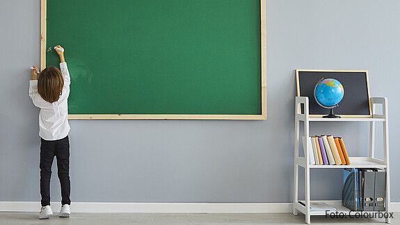 Das foto zeigt ein Kind, das eine Tafel beschriftet. Rechts ist ein Regal mit einem Globus zu sehen.