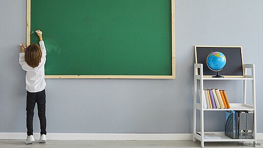 Das foto zeigt ein Kind, das eine Tafel beschriftet. Rechts ist ein Regal mit einem Globus zu sehen.