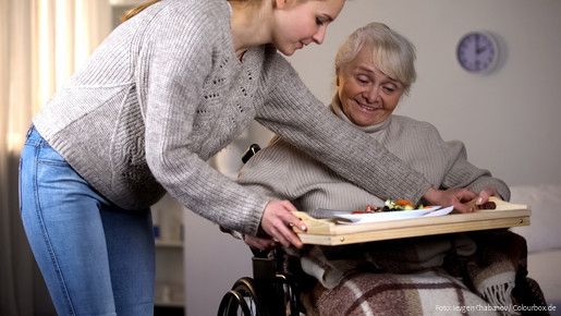 Eine junge Frau reicht einer älteren Frau im Rollstuhl ein Tablett mit einem Mittagessen.