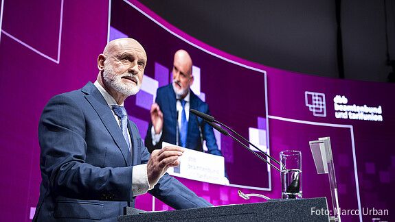 Ein Mann steht auf einer Bühne hinter einem Rednerpult und spricht in ein Mikrofon. 