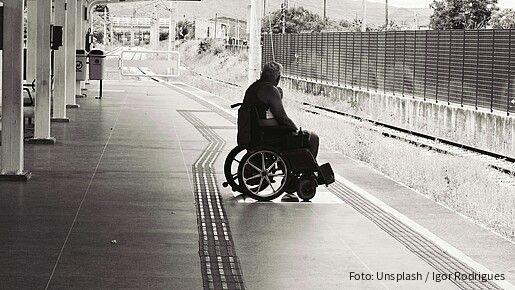Schwarz-weiß-Foto eines Mannes, der an einem Bahnsteig in einem Rollstuhl sitzt und auf den Zug wartet.