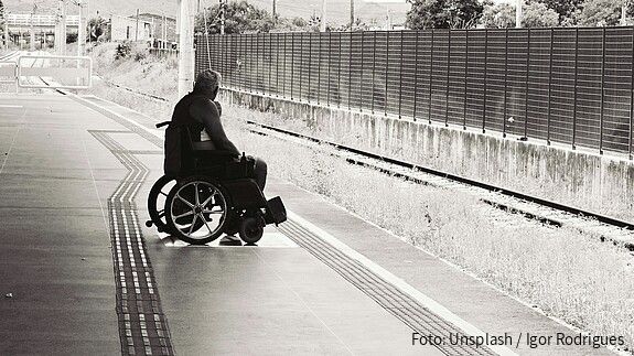 Schwarz-weiß-Foto eines Mannes, der an einem Bahnsteig in einem Rollstuhl sitzt und auf den Zug wartet.