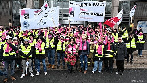Eine Gruppe Menschen demonstriert mit bunten Bannern und Fahnen auf den Stufen eines Krankenhauses
