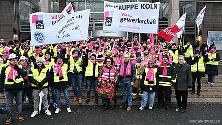 Eine Gruppe Menschen demonstriert mit bunten Bannern und Fahnen auf den Stufen eines Krankenhauses