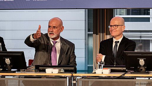 Zwei Männer in Anzügen sitzen an einem Tisch uns sprechen in ein Mikrofon