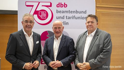 Drei Männer in Anzügen stehen vor einem Aufsteller und blicken in die Kamera