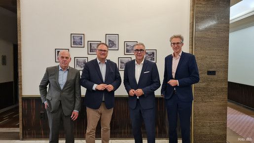 Vier Männer in Anzügen stehen vor einer Wand mit Bildern und blicken in die Kamera