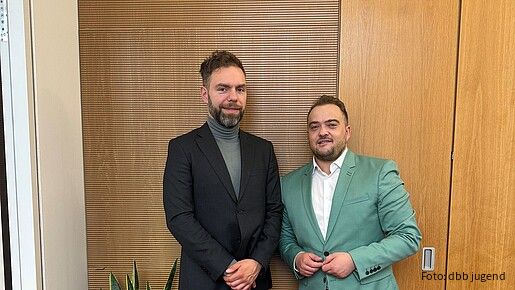 Zwei Männer in Anzügen stehen vor einer Holzwand und blicken in die Kamera